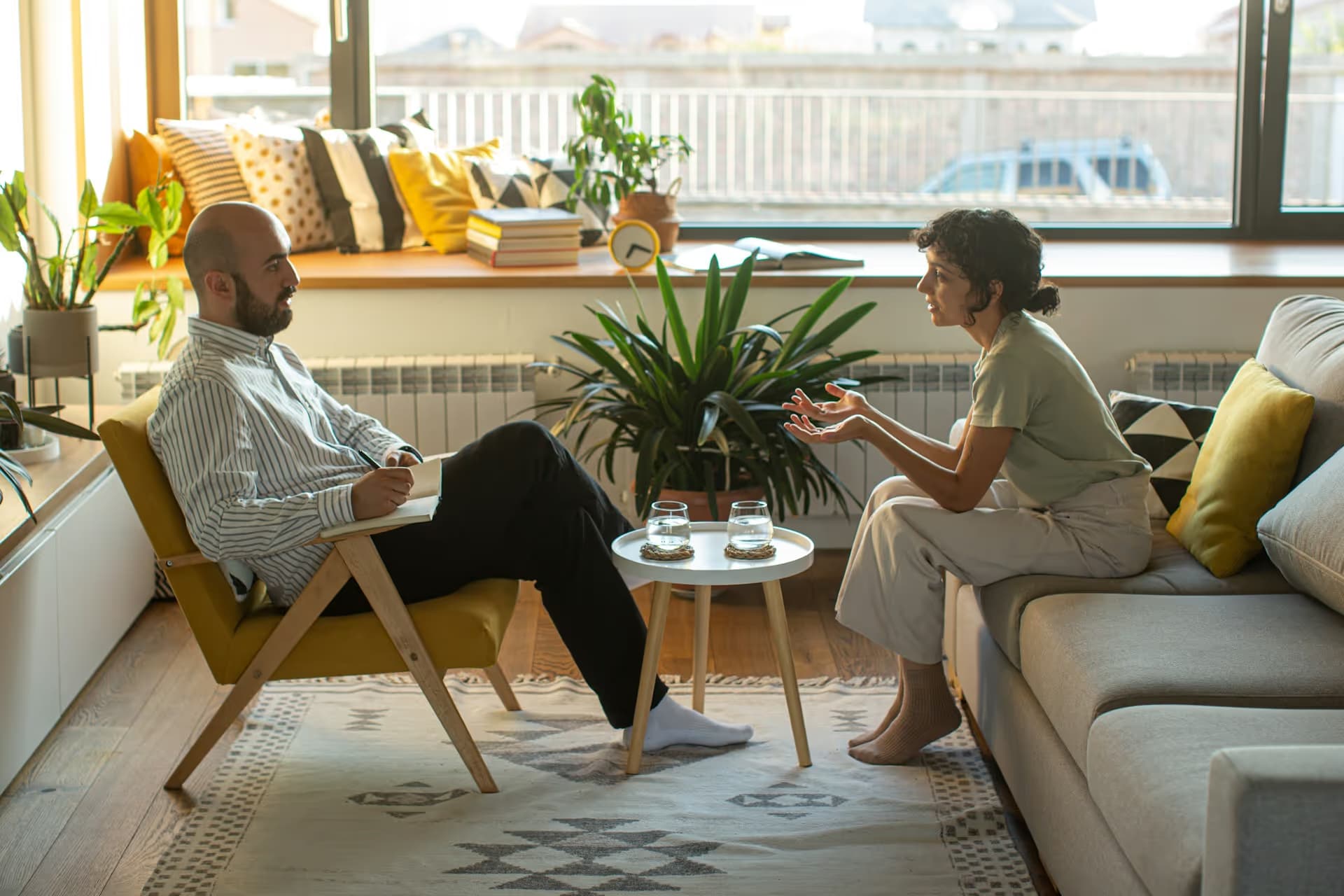 A man in a striped shirt sitting on a yellow chair with a notebook, listening to a woman in casual clothes seated on a gray sofa talking animatedly.