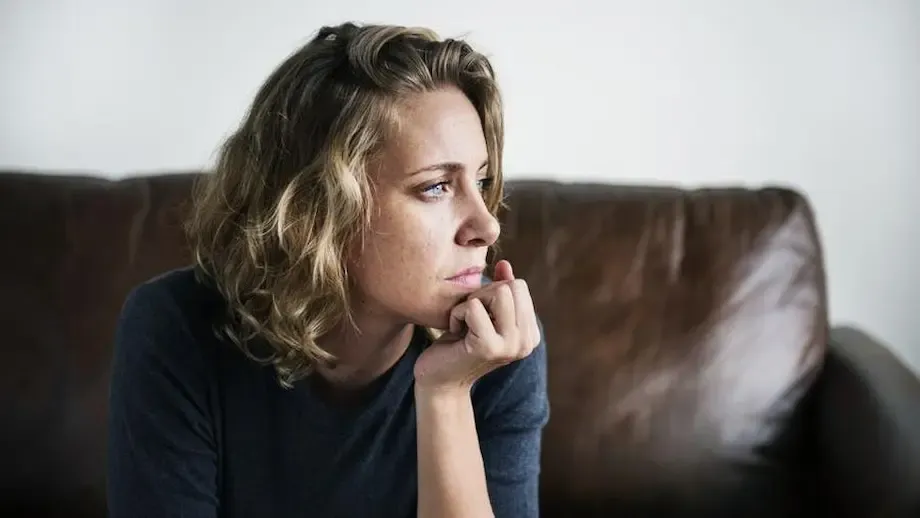 A woman with blonde curly hair, wearing a dark shirt, sitting on a brown leather couch, looking pensively to her left with her chin resting on her hand.