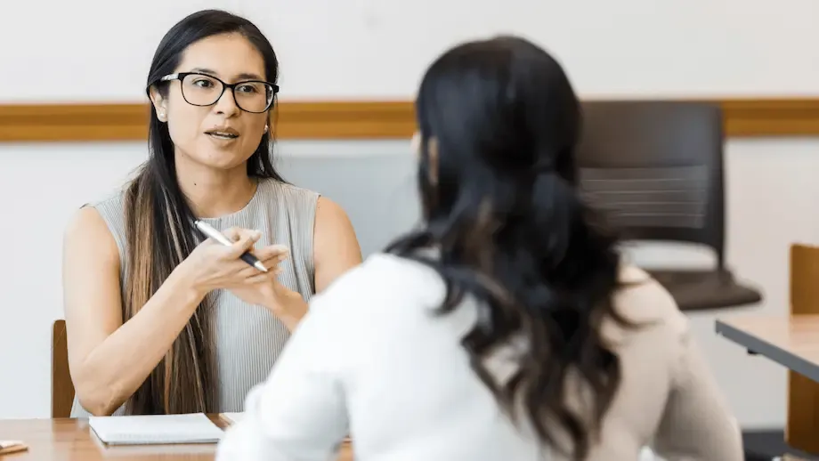 Two women seated at a table engaged in a conversation. The woman facing the camera, wearing glasses and a sleeveless top, is speaking and gesturing with a pen, while the other woman, with her back to the camera, listens attentively. The setting appears to be a professional or educational environment.