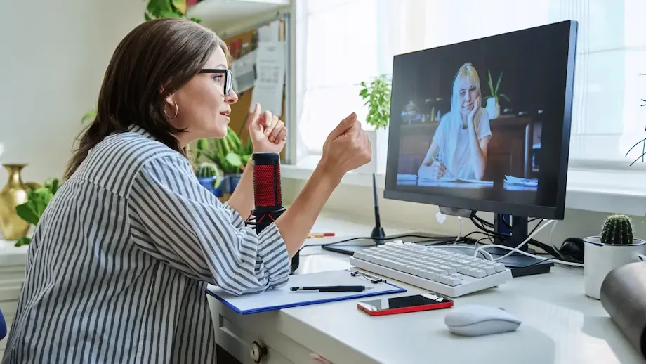 A woman sitting at a desk, engaged in a video call with another woman displayed on the computer screen. The woman at the desk is speaking and gesturing with her hands, surrounded by various office items and plants