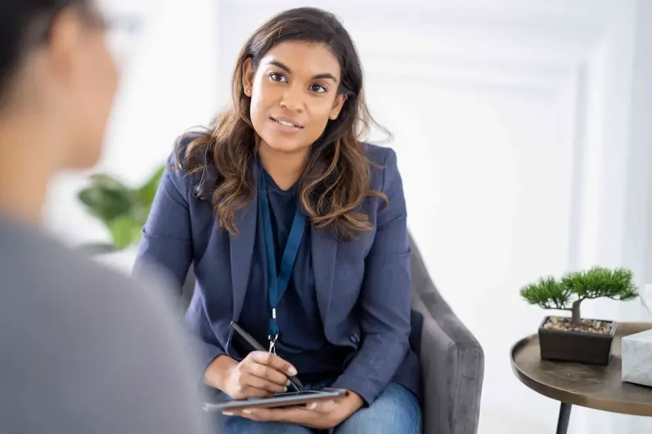 A female psychiatrist or therapist sitting in an office, attentively listening to a patient who is partially visible in the foreground. She is holding a clipboard and a pen, dressed in a blazer and casual attire, with a small bonsai tree on a table nearby.