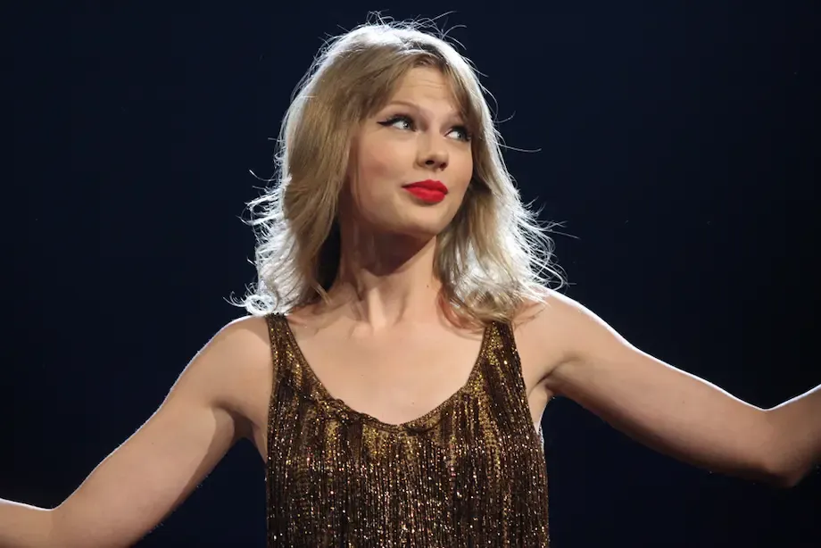 A famous female singer with blonde hair wearing a glittery gold dress, standing on stage with her arms outstretched and a confident expression, under bright stage lighting