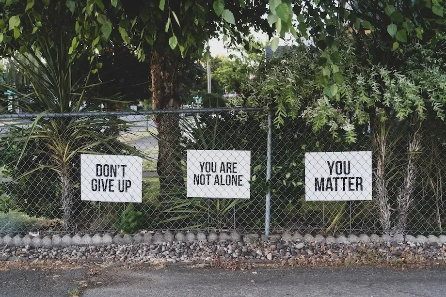 A chain-link fence with three motivational signs attached, reading 'DON'T GIVE UP', 'YOU ARE NOT ALONE', and 'YOU MATTER'. The fence is surrounded by greenery and trees.
