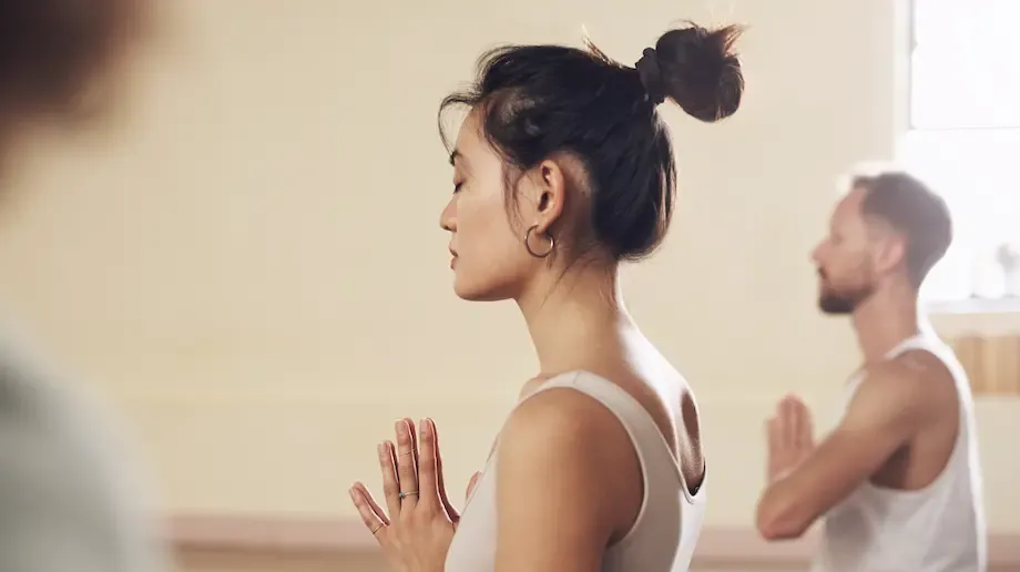 A woman in a yoga class, standing with hands in prayer position, eyes closed, focusing inward. She is in a peaceful, well-lit studio with other participants visible in the background.