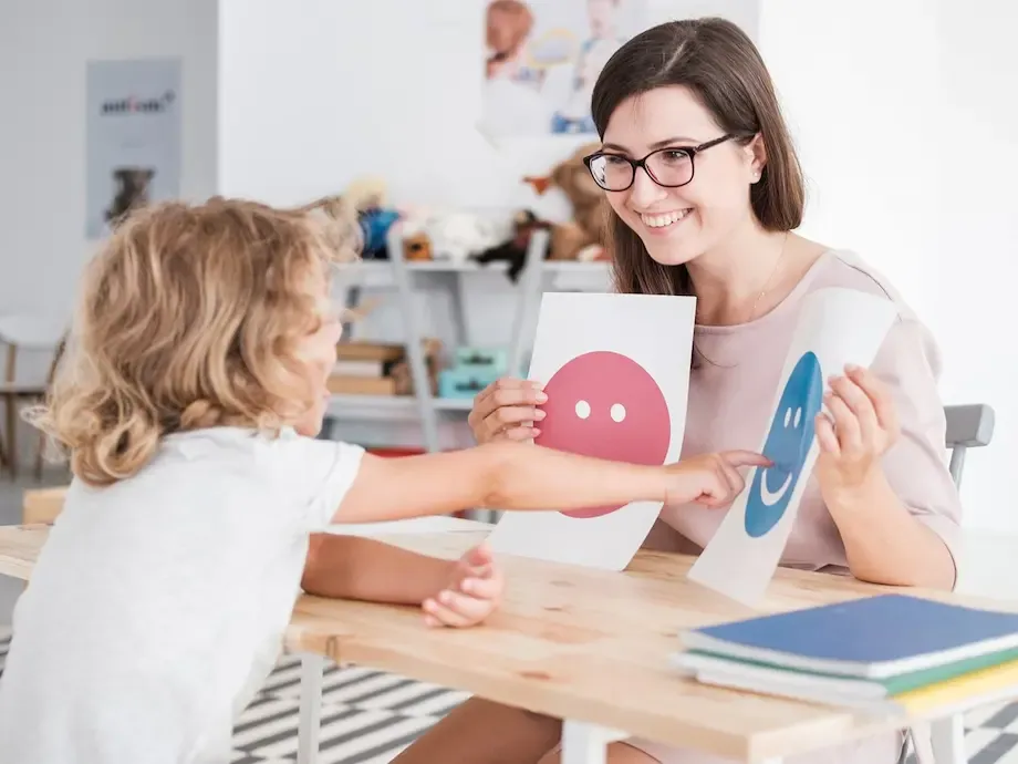 A young child and a therapist sitting at a table in a bright and friendly room. The therapist, a woman with glasses, is smiling and holding up a card with a happy face, while the child reaches for a card with a sad face, illustrating an emotional recognition exercise