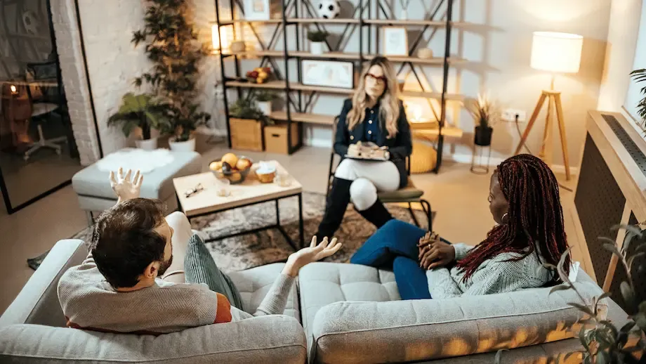 A therapy session in progress in a warmly lit, stylish living room. Two clients, a man and a woman, are seated on separate gray sofas, gesturing while speaking animatedly. The therapist, a woman in glasses, listens intently while seated on a chair across from them, creating an environment of open communication and support
