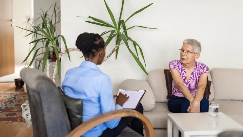 An elderly woman sitting on a sofa during a therapy session, discussing with a therapist who is taking notes. The room is comfortably furnished, with houseplants adding a touch of nature