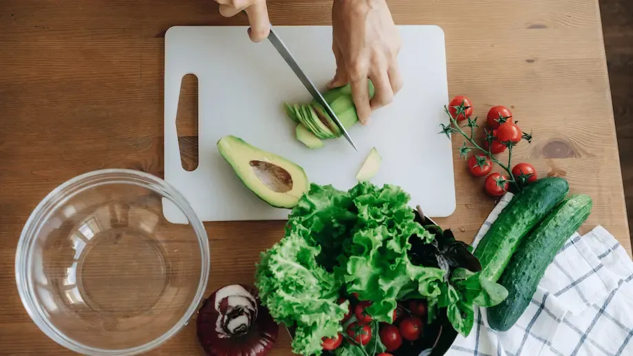 An overhead view of a wooden kitchen counter where someone is slicing an avocado next to a bowl and other fresh vegetables like lettuce, tomatoes, and cucumbers, preparing a healthy salad.