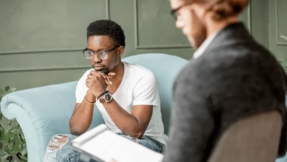A young African American man thoughtfully listens to a therapist who is sitting across from him, holding a clipboard in a softly lit room.