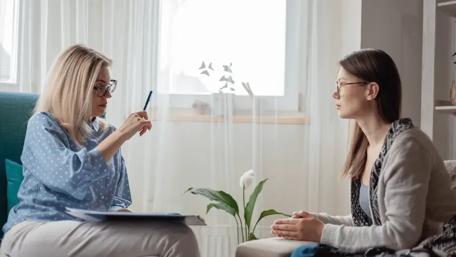 A calm therapy session featuring a female therapist and a young woman sitting across from each other, engaging in a serious conversation.