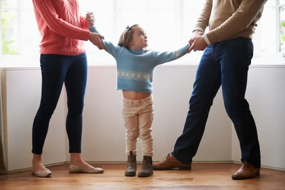 A young child trying to hold hands with both parents in a bright indoor setting, symbolizing a moment of family unity or mediation, as the parents stand apart with concerned expressions