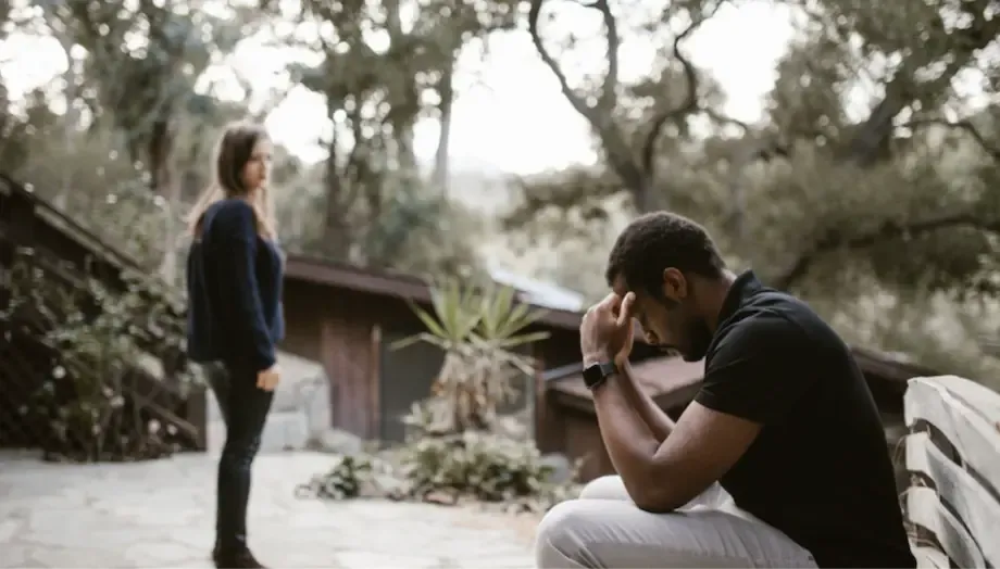 A tense scene outdoors where a man sits with his head in his hands, looking distressed, while a woman stands a distance away looking back at him, both appearing to be in a serious conversation or disagreement in a wooded area.