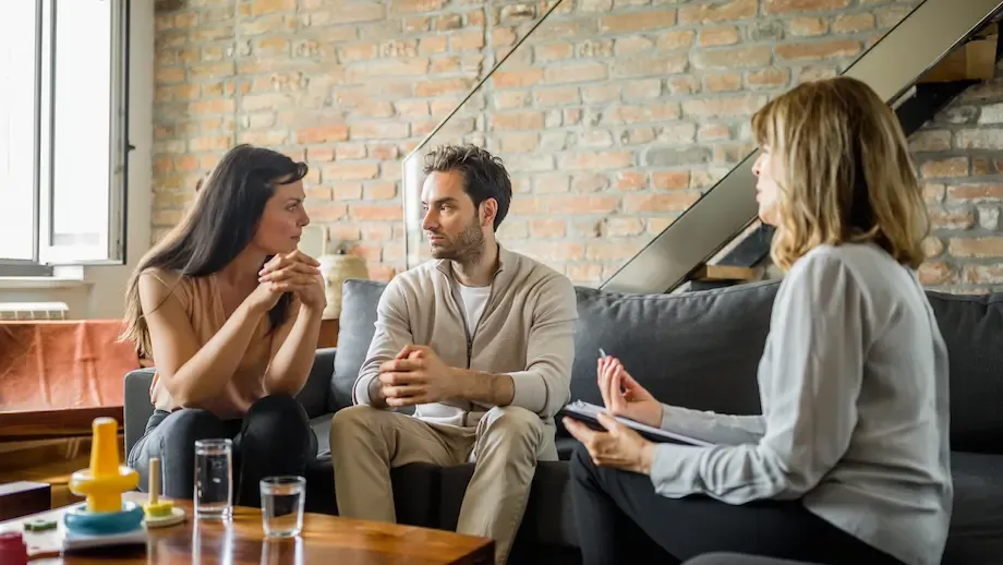 A family therapy session showing a young African American couple sitting together on a sofa, smiling and talking to their young daughter who stands between them, while a therapist sits nearby taking notes.