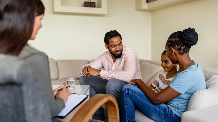 A young couple sits on a sofa during a therapy session, holding hands and looking intently at each other as they talk, with a female therapist sitting across from them, holding a clipboard.