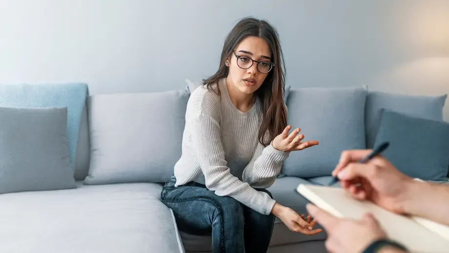 A young woman in a light sweater sits on a gray sofa, engaged in a conversation with her therapist, who is taking notes. Her expression is earnest and slightly concerned.