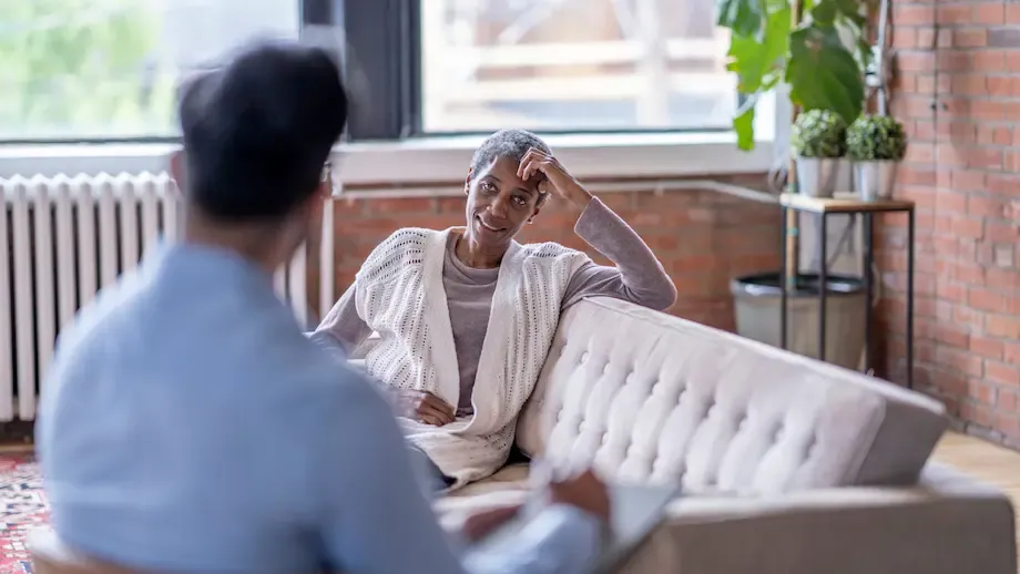 A senior African American woman in a cozy sweater sits on a sofa in a well-lit therapy room, looking thoughtful while talking to her therapist, whose back is to the camera.