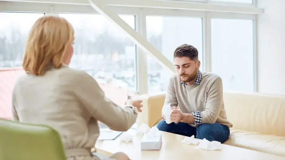 A bright therapy room with large windows showing a snowy exterior. A male client, looking contemplative and slightly distressed, sits on a couch discussing his feelings with a female therapist who is listening intently