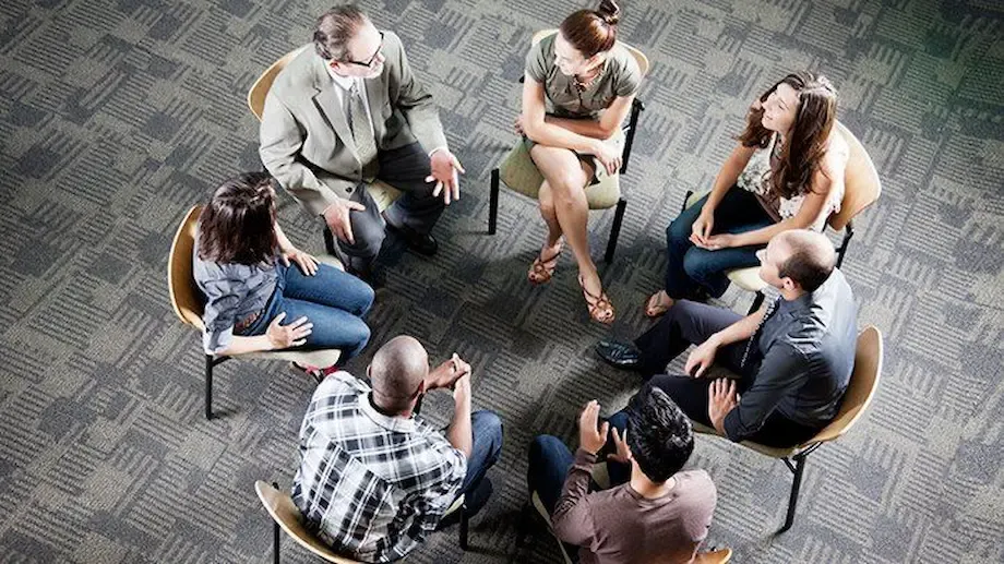A diverse group of eight adults seated in a circle on chairs during a group therapy session. They are engaged in a discussion led by a therapist, with participants expressing themselves and listening attentively in a casual, supportive setting.