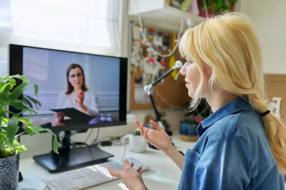 A woman participates in a video call therapy session, speaking with a female therapist displayed on her computer screen. The woman, with blonde hair, gestures with one hand while seated at a desk in a brightly lit, creative workspace. The therapist, shown on screen, appears attentive and engaged, sitting in a professional setting