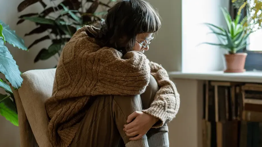 A young woman, wrapped in a chunky knit sweater, sits curled up on an armchair by a window with a view of green plants, appearing contemplative and sad