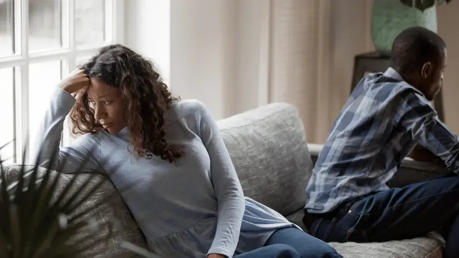 An African-American couple experiencing tension, seated apart on a couch. The woman, dressed in a light blue sweater, looks distressed and rubs her forehead, while the man, in a blue plaid shirt, looks away with an expression of concern.