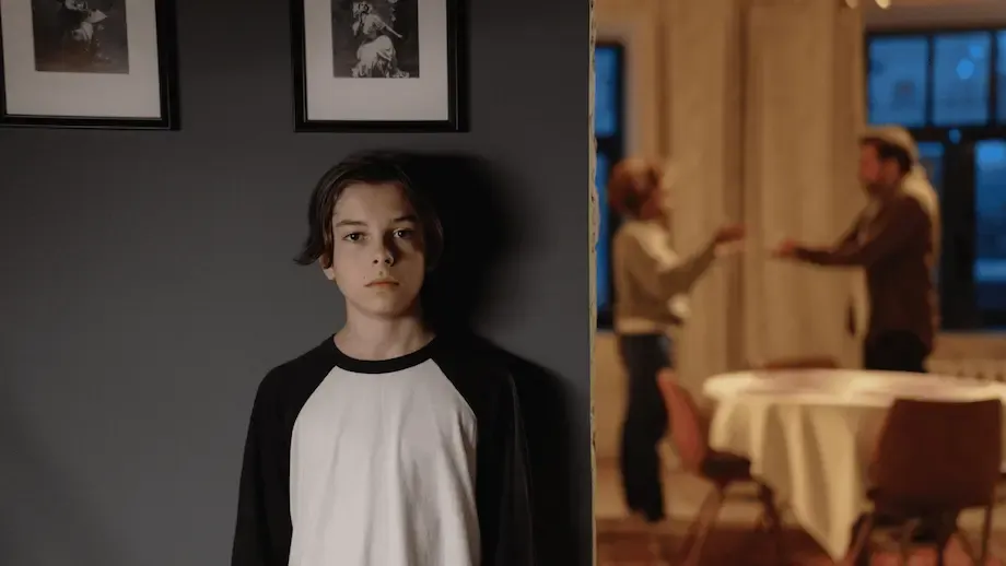 An image split into two scenes: On the left, a young boy in a black and white baseball tee stands solemnly against a dark wall decorated with framed photos. On the right, two adults, a man and a woman, appear in a heated discussion in a warmly lit dining area