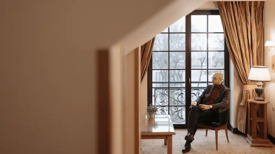 An older bald man dressed in a dark suit and glasses, sitting contemplatively in an elegantly furnished room with large windows covered by heavy drapes, looking out onto a snowy landscape