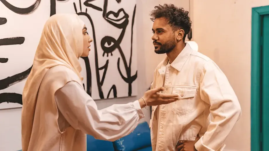 A Middle Eastern man and woman having a tense conversation in an art gallery. The woman, wearing a hijab, gestures with her hand while looking seriously at the man, who is wearing a beige shirt and listens with a slight frown