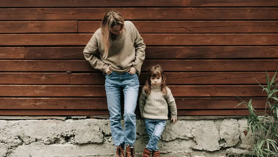 A Caucasian mother and daughter standing next to each other against a wooden wall, both dressed in warm clothing. The mother is bending over slightly towards the daughter, who appears shy or hesitant.