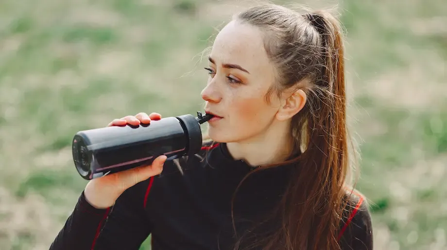 A woman drinking from a sports water bottle while taking a break outdoors. She has long brown hair, is dressed in sportswear, and looks refreshed, with a grassy field in the background