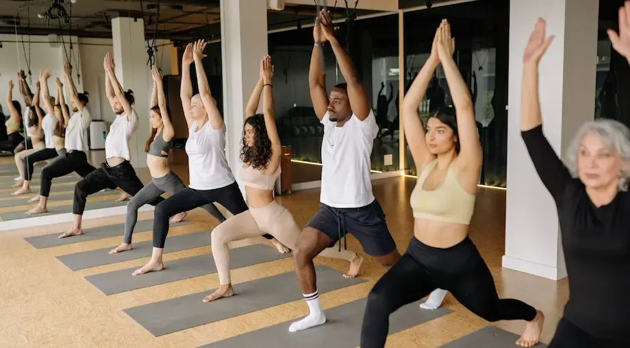 A diverse group of people participating in a yoga class, performing a standing yoga pose with their arms raised. The room is spacious and brightly lit, with yoga mats aligned on the floor