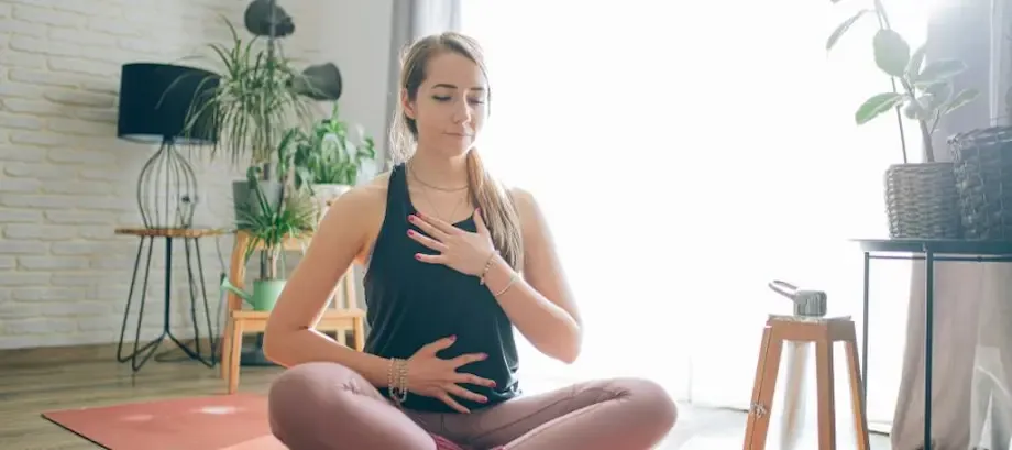 A woman seated cross-legged on a yoga mat in her living room, practicing meditation. She has her hands on her knees, eyes closed, with a serene expression, surrounded by indoor plants.