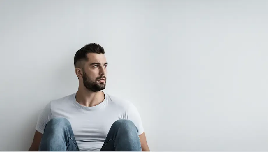 A young man sitting on the floor against a plain white background, wearing a white t-shirt and jeans. He looks off to the side thoughtfully, with a serious expression