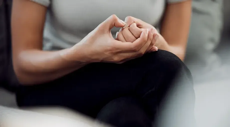 A close-up of a man's hands, anxiously clasped together on his lap, as he sits on a couch. His arms are muscular, and his posture suggests tension or nervousness
