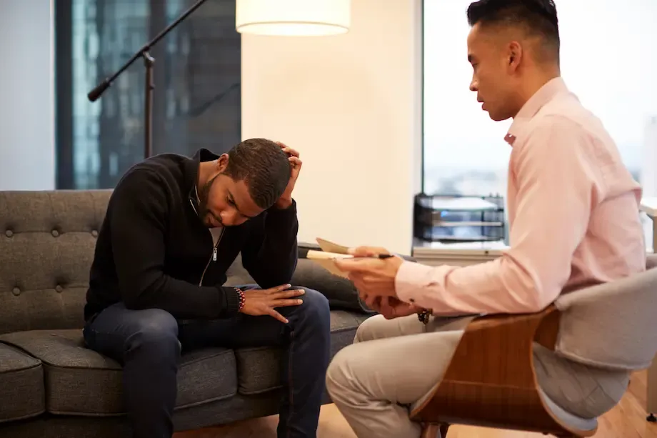 A counseling session in a modern office setting where a distressed man, seated on a dark gray sofa, holds his head with one hand, showing signs of emotional stress. Opposite him, a therapist in a light pink shirt and gray pants sits on a wooden chair, holding a notepad and listening attentively.