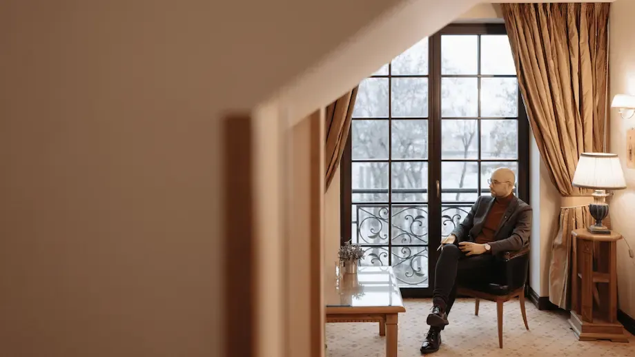 An elegantly furnished room with a well-dressed bald man sitting on a chair, looking thoughtful. The room features stylish drapes and a French window overlooking a snowy landscape, giving the scene a serene and contemplative atmosphere