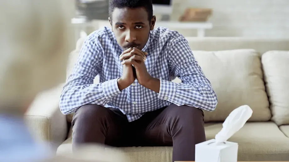 A focused African American man sitting on a sofa in a bright living room, with his hands clasped and a serious expression on his face. He seems to be deep in thought or waiting for someone to speak, with a box of tissues placed next to him on the sofa.