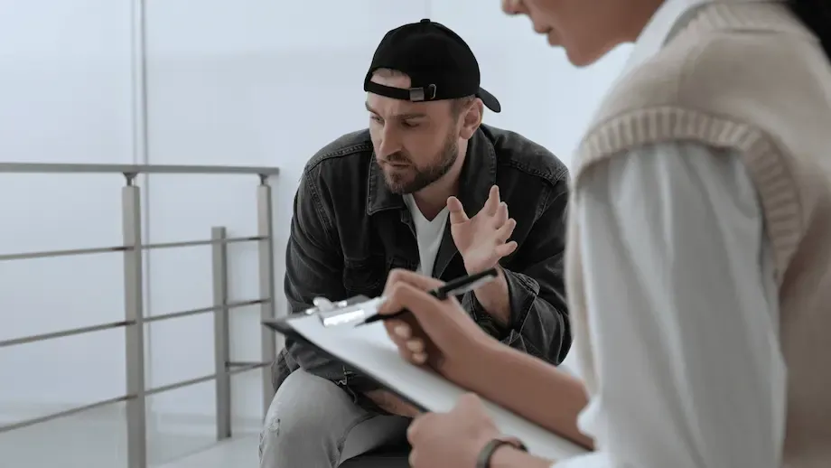 A therapy session in progress with a male patient in distress, seated on a couch, discussing his issues with a therapist who is attentively taking notes. The patient, wearing a dark jacket and a cap, appears troubled as he talks, gesturing with his hand.
