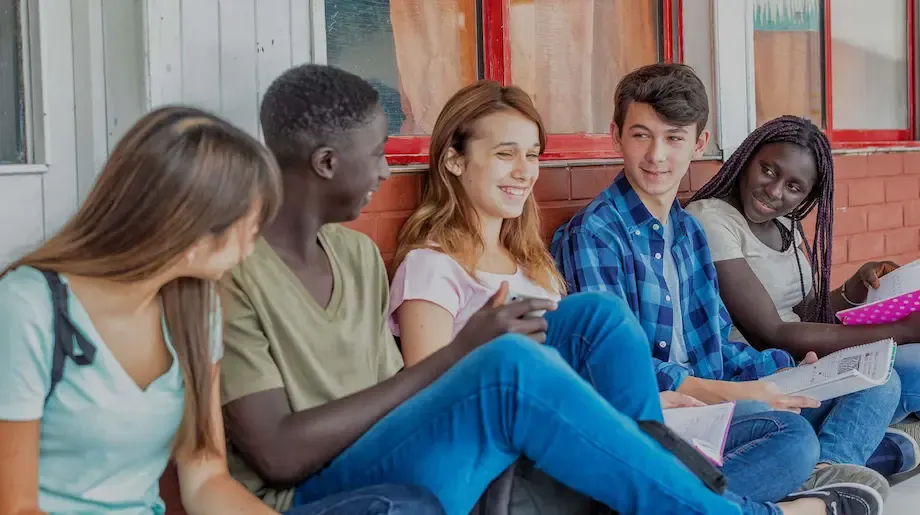 A group of five diverse teenagers sitting and chatting on a school bench. From left to right, a Hispanic girl, a Black boy, a Caucasian girl holding a smartphone, an Asian boy, and a Black girl with braids. They are all casually dressed, holding books and backpacks, laughing and engaging in a friendly conversation in a relaxed outdoor school setting