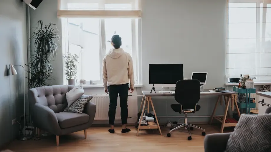 A rear view of a person standing in a spacious and stylishly furnished modern living room, looking out of a large window. The room is equipped with a contemporary sofa and a home office setup, suggesting a peaceful, comfortable living space