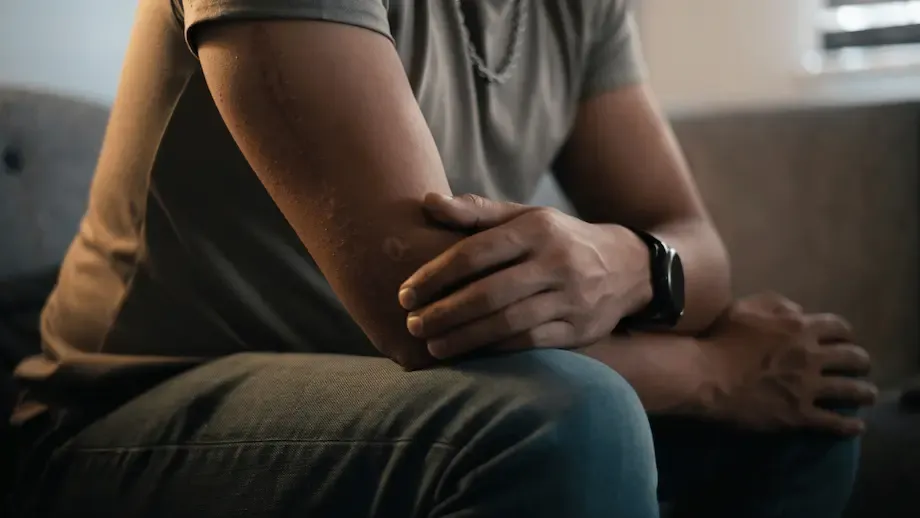 A close-up of a man's hands clasped together while sitting, showing signs of worry or contemplation. The focus on his hands and the blurred background emphasize a moment of anxiety or deep thought