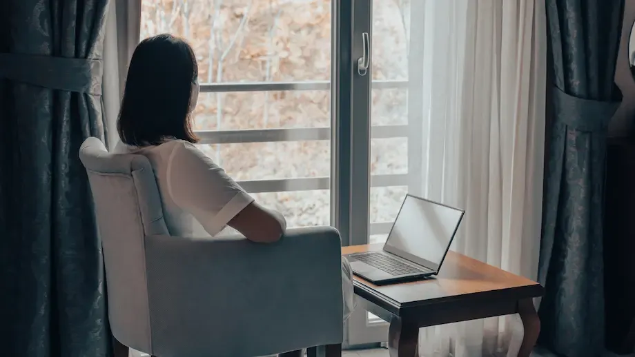 A woman sits in a modern, well-lit office, looking out a window thoughtfully. She is dressed in a casual white shirt, with her laptop open on the desk beside her, suggesting a break or a moment of reflection during her workday