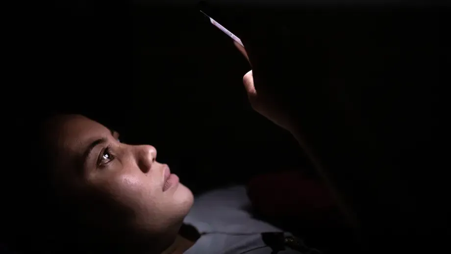 A young adult lies in bed at night, staring intently at their smartphone screen, which illuminates their face in the otherwise dark room, capturing a moment of solitude and digital engagement