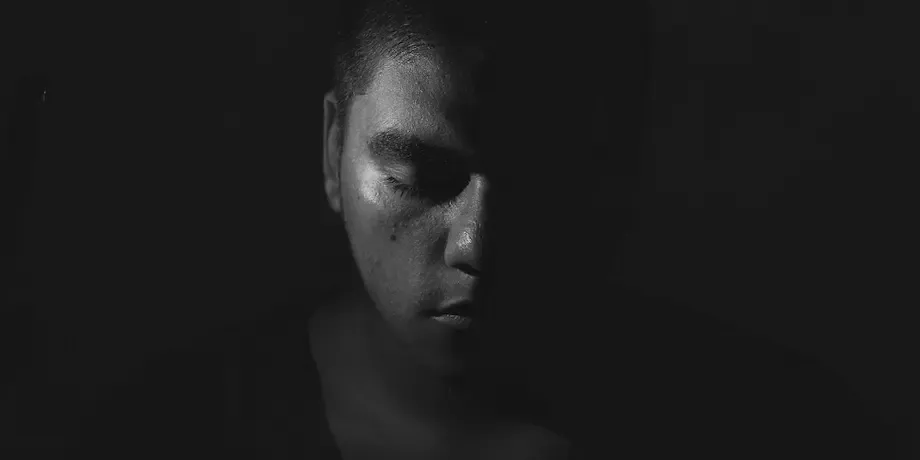 A striking black and white portrait of a man in profile, his face partially illuminated and casting a somber expression. The dramatic lighting accentuates the contours of his face and the texture of his skin, conveying a mood of introspection or melancholy.