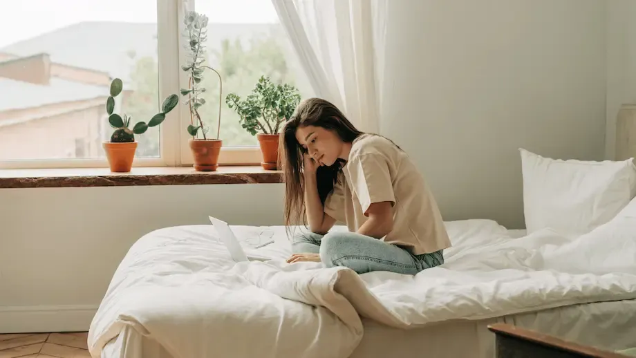 Young woman sitting cross-legged on her bed, gazing thoughtfully at papers in front of her, with sunlight illuminating the room and plants by the window.