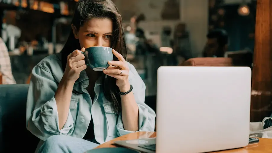 Woman in a light denim shirt sips coffee while looking at a laptop in a cozy cafe setting, her focused expression suggesting engagement with her work