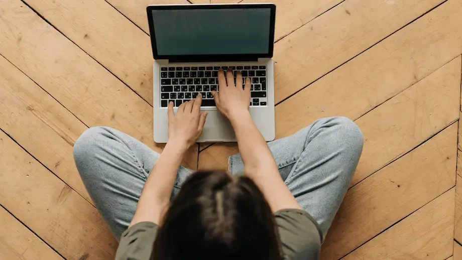 Aerial view of a woman seated on a hardwood floor typing on a laptop, capturing her relaxed posture and casual home environment