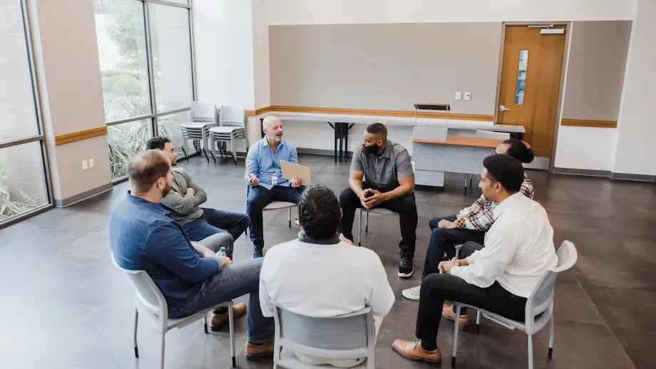 A group therapy session featuring a diverse group of men sitting in a circle inside a spacious room with large windows, engaging in a conversation led by a therapist dressed in a blue shirt and jeans