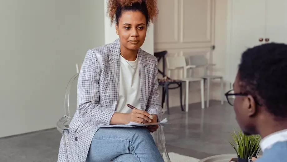 A therapist, a young woman with curly hair and a plaid blazer, sits on a clear plastic chair, attentively taking notes during a session with a male client, who is partially visible and appears to be discussing his concerns.