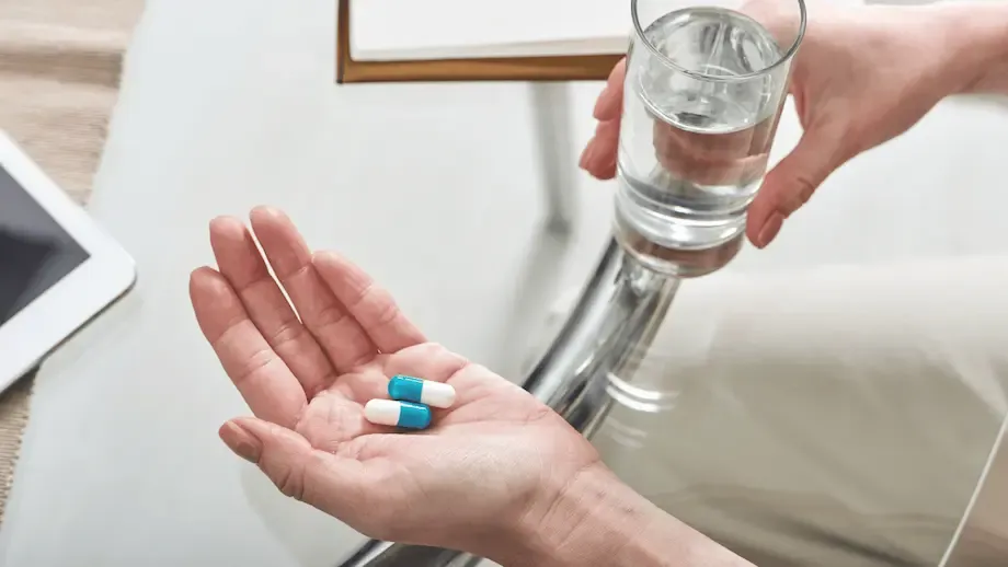 A close-up of a person's hands holding two blue and white capsules in one hand and a glass of water in the other, ready to take medication. A tablet device is visible on the table next to them